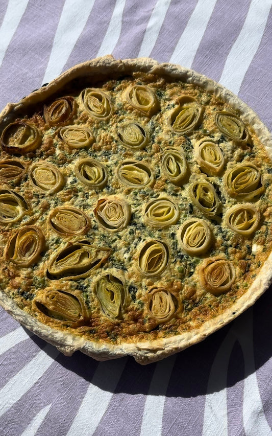 Puss pastry tart filled with spinach, peas and carmelised leeks that have been cut into discs and placed on the top of the tart. Tart has been baked and is brown on the edges and is sitting on top of a lilac zebra print table cloth.